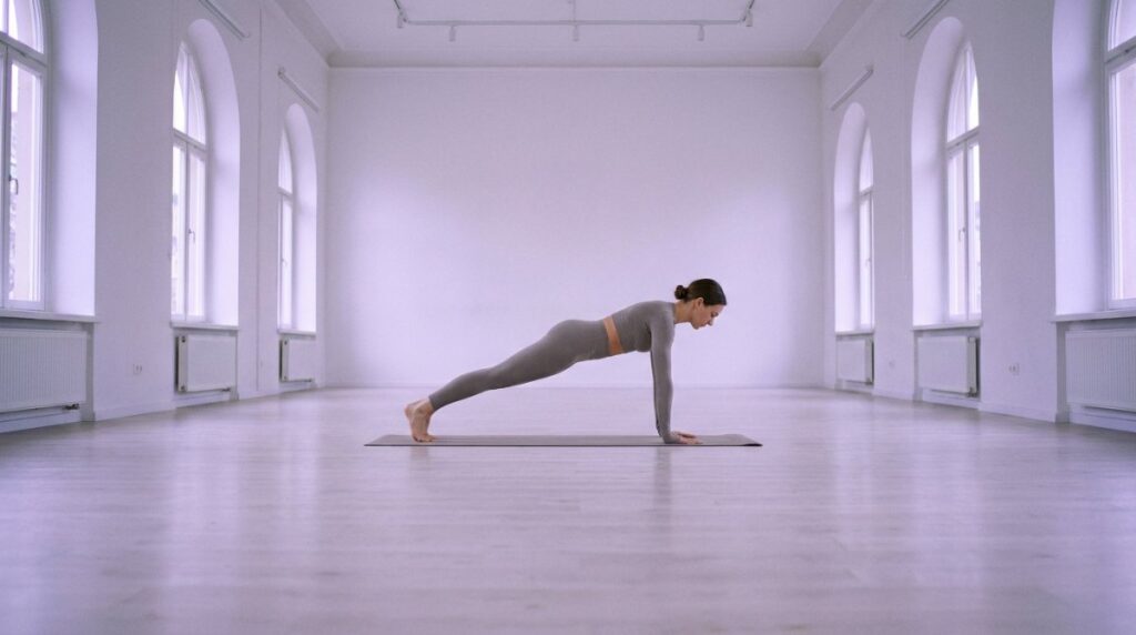 A woman holding a controlled plank position on a mat in a spacious light-filled Pilates studio